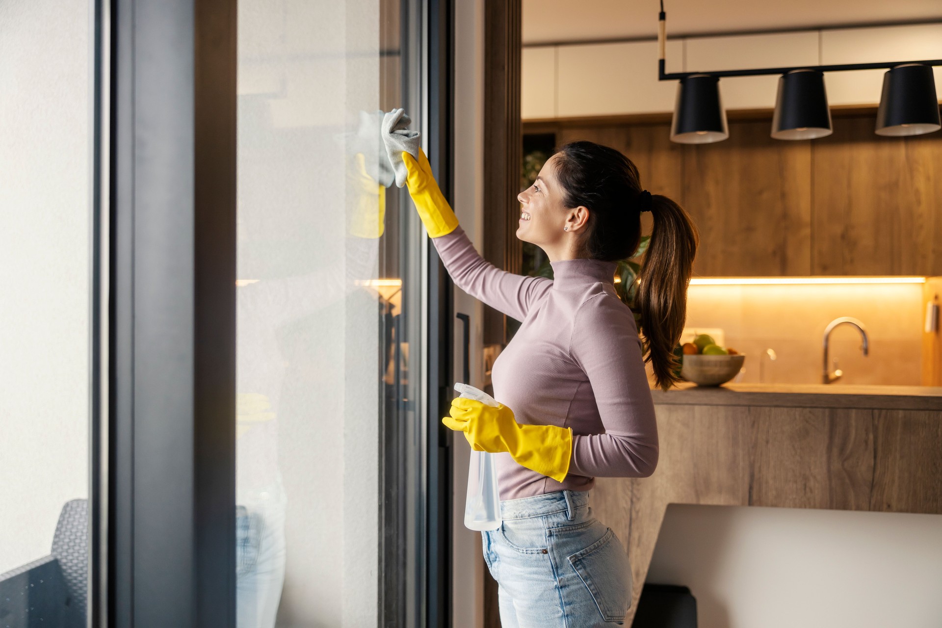 Neat young woman with ribber gloves cleaning windows at home with detergent and rag.